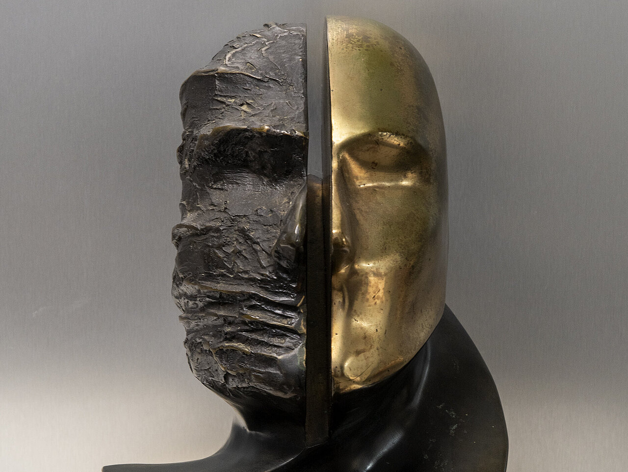 Die Bronzeskulptur zeigt ein geteiltes Kopfporträt, wobei eine Hälfte rau und die andere glatt und vergoldet ist.