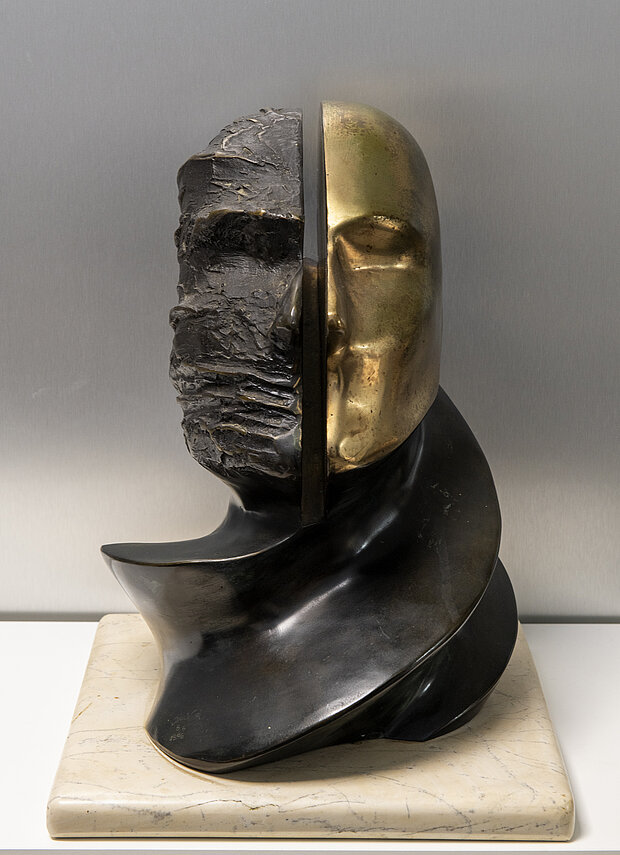 Die Bronzeskulptur zeigt ein geteiltes Kopfporträt, wobei eine Hälfte rau und die andere glatt und vergoldet ist.